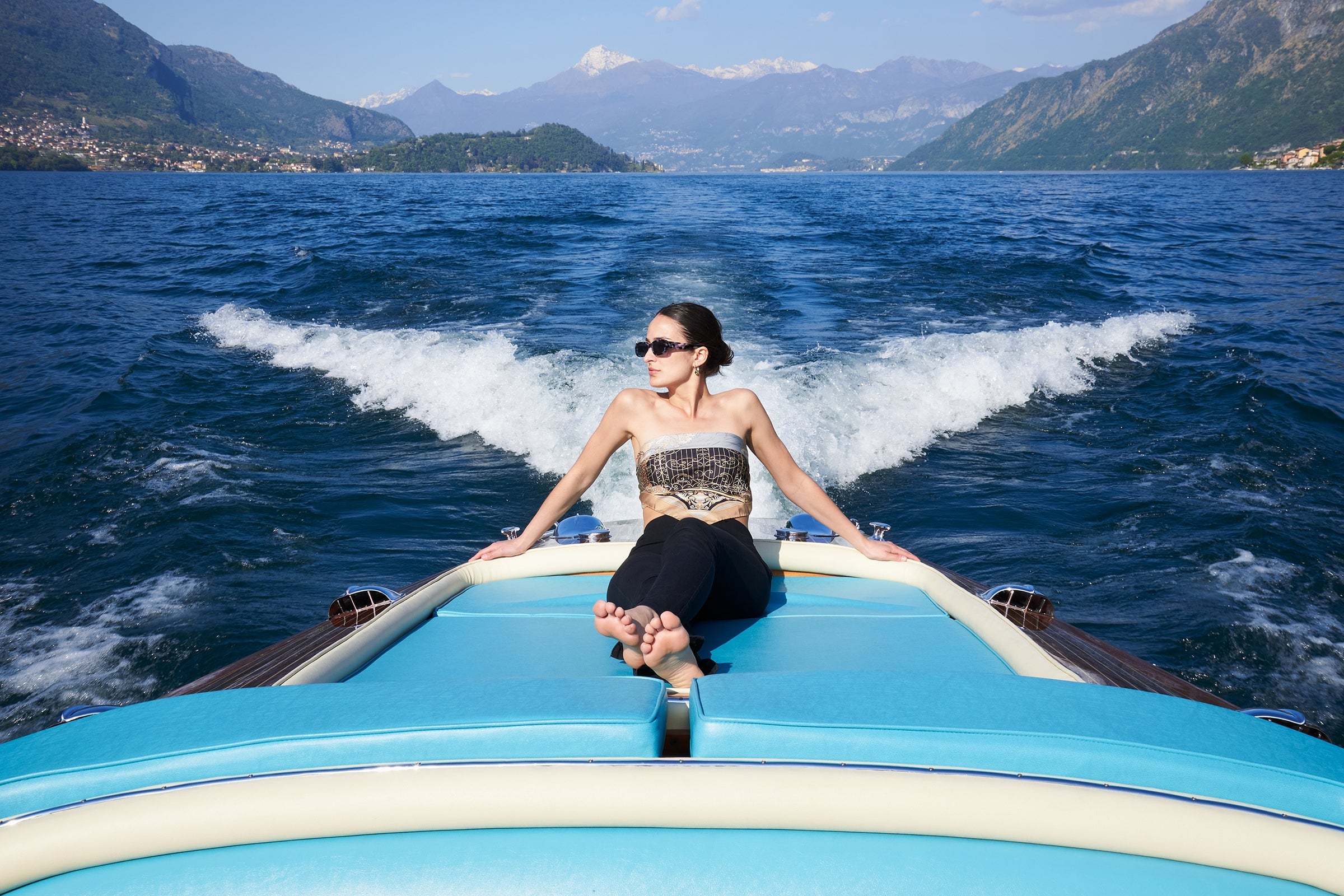 woman wearing sunglusses and a silk scarf as shirt, sitting on a motorboat on the waters of Lake Como. On the background the lake, Tremezzina, Lenno, Villa Balbianello and the mountains